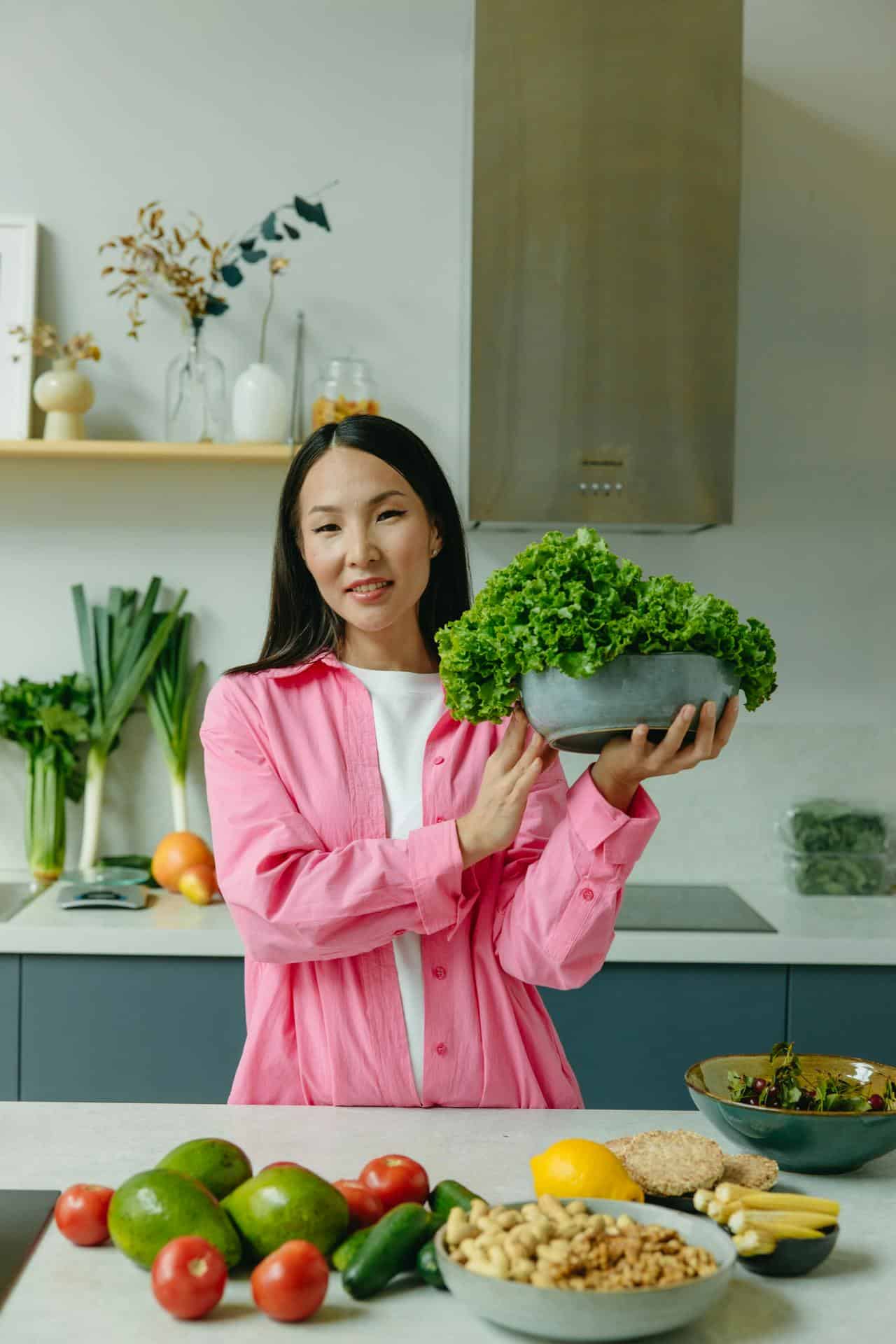 Mesma mulher de rosa, agora segurando uma bacia de couve com alguns legumes na mesa.